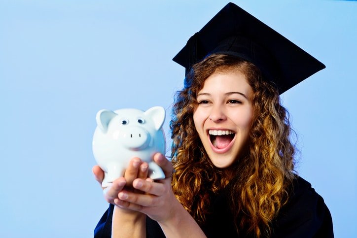 Delighted young graduate in academic dress holds up piggybank