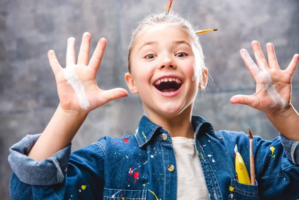 Schoolgirl artist with painted face showing palms in paint and smiling at camera